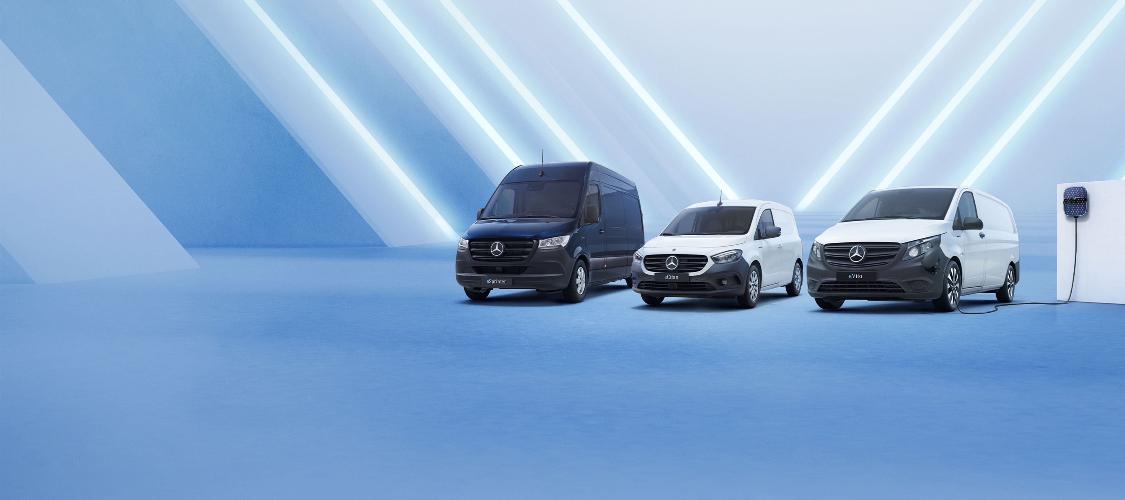 Elektriske varebiler | Mercedes-Benz Vans Tre elektriske Mercedes-Benz varebiler på række ved ladestander på væggen til højre