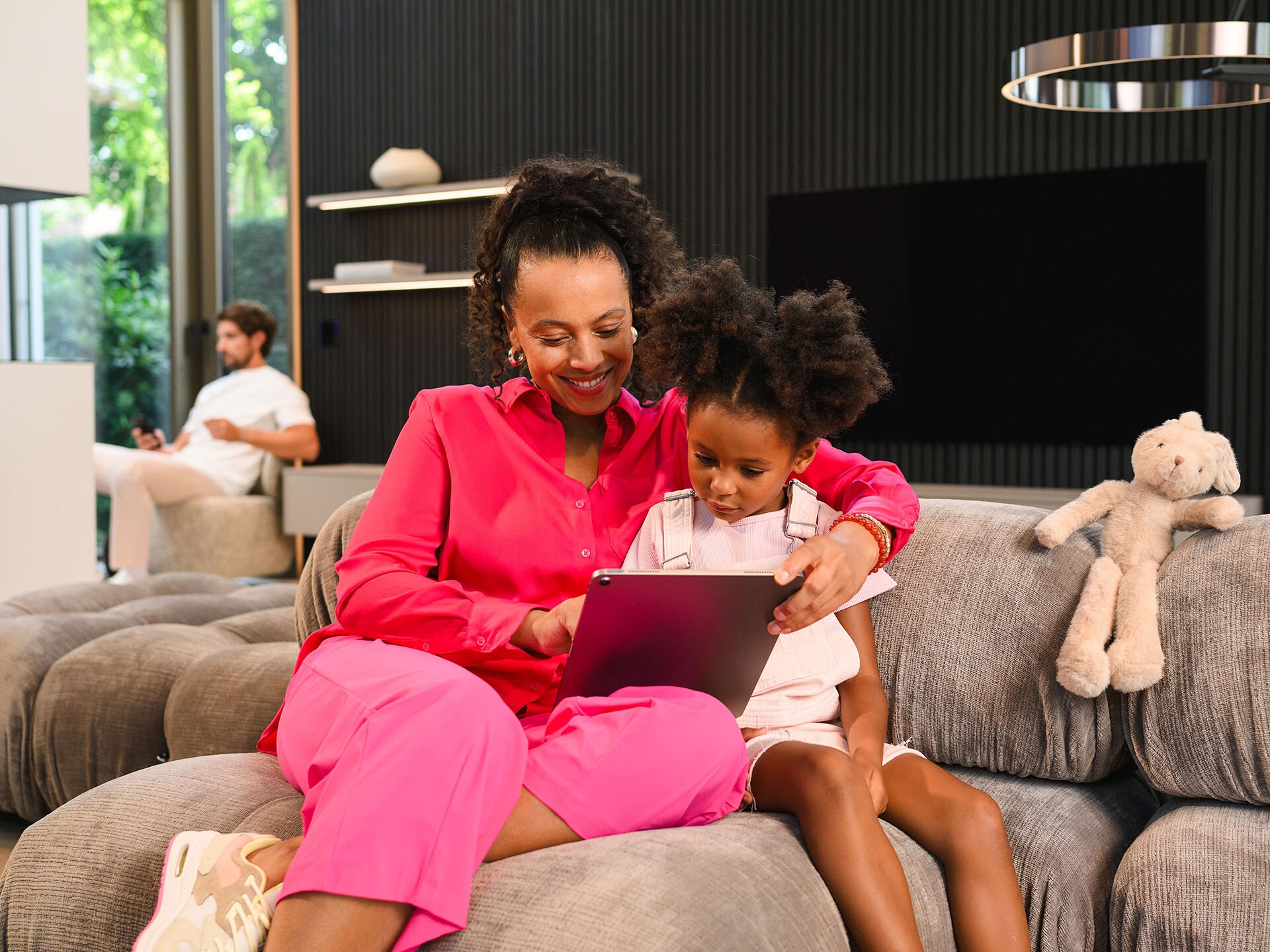 En kvinde og et barn, der sidder i en sofa og bruger en tablet, med en mand der sidder på baggrunden.