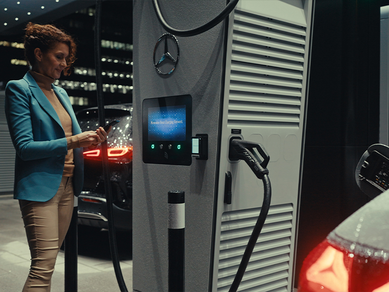 Mercedes-Benz app | Opladning undervejs | Mercedes-Benz En kvinde med en smartphone i hånden, der står ved siden af en ladestation.