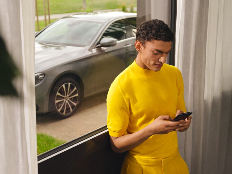 Mand med smartphone står ved vindue ud mod indkørslen, hvor bilen er parkeret