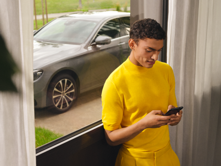 Mercedes-Benz Finans | Kundeservice | Mercedes-Benz Mand med smartphone står ved vindue ud mod indkørslen, hvor bilen er parkeret