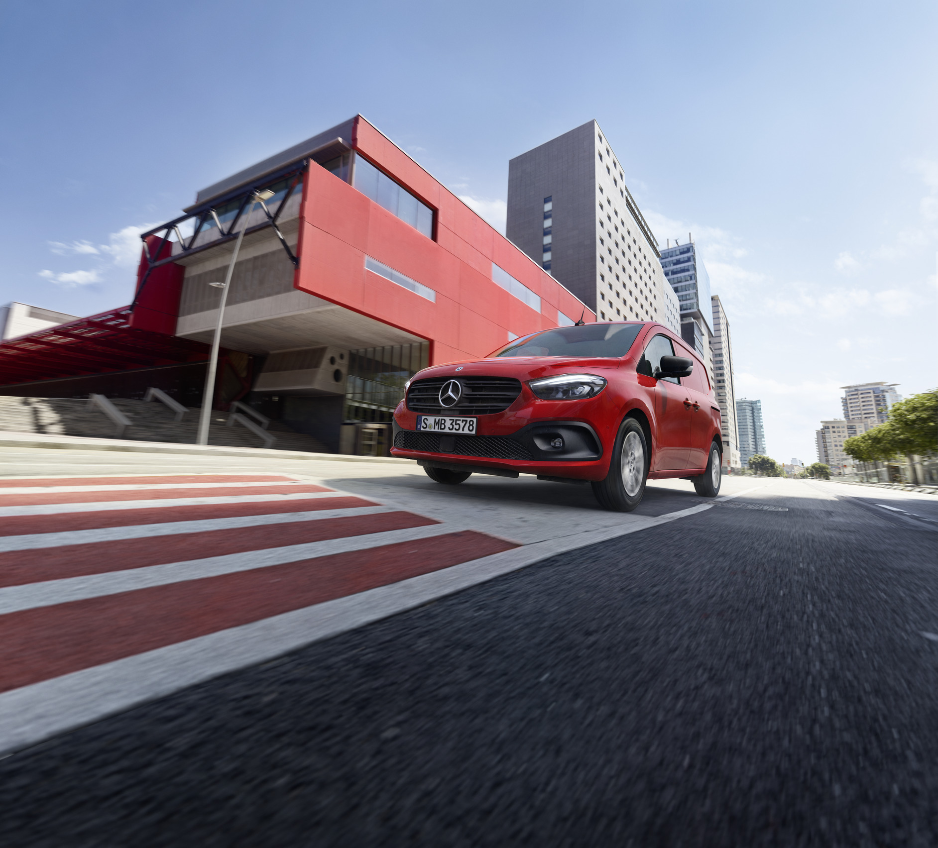 eCitan og Citan | Mercedes-Benz Vans En rød Mercedes-Benz Citan Kassevogn på farten med rød bygning i baggrunden