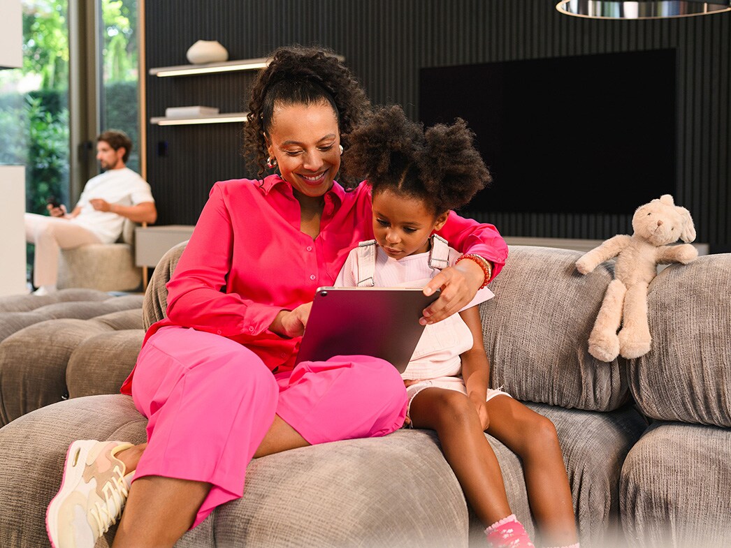 En kvinde og et barn, der sidder i en sofa og bruger en tablet, med en mand der sidder på baggrunden.