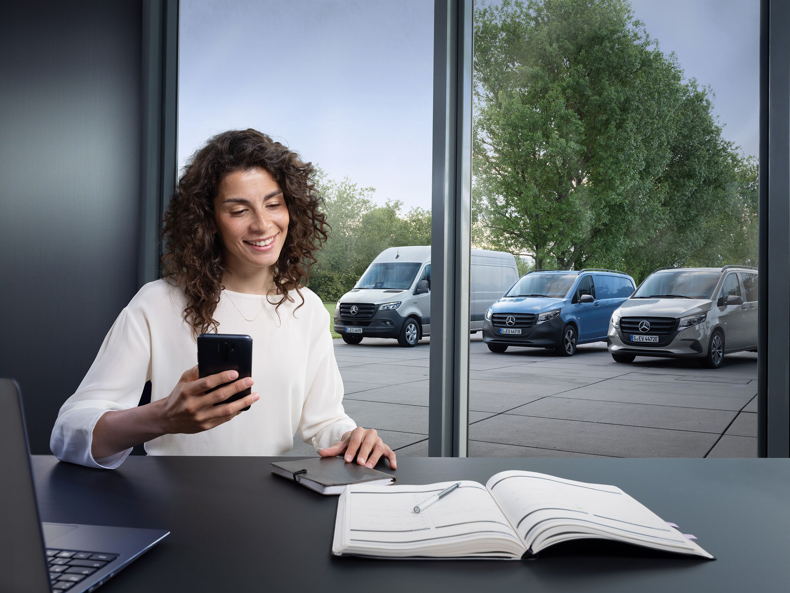 Kvinde med smartphone ved et skrivebord, flere Mercedes-Benz varebiler i baggrunden.