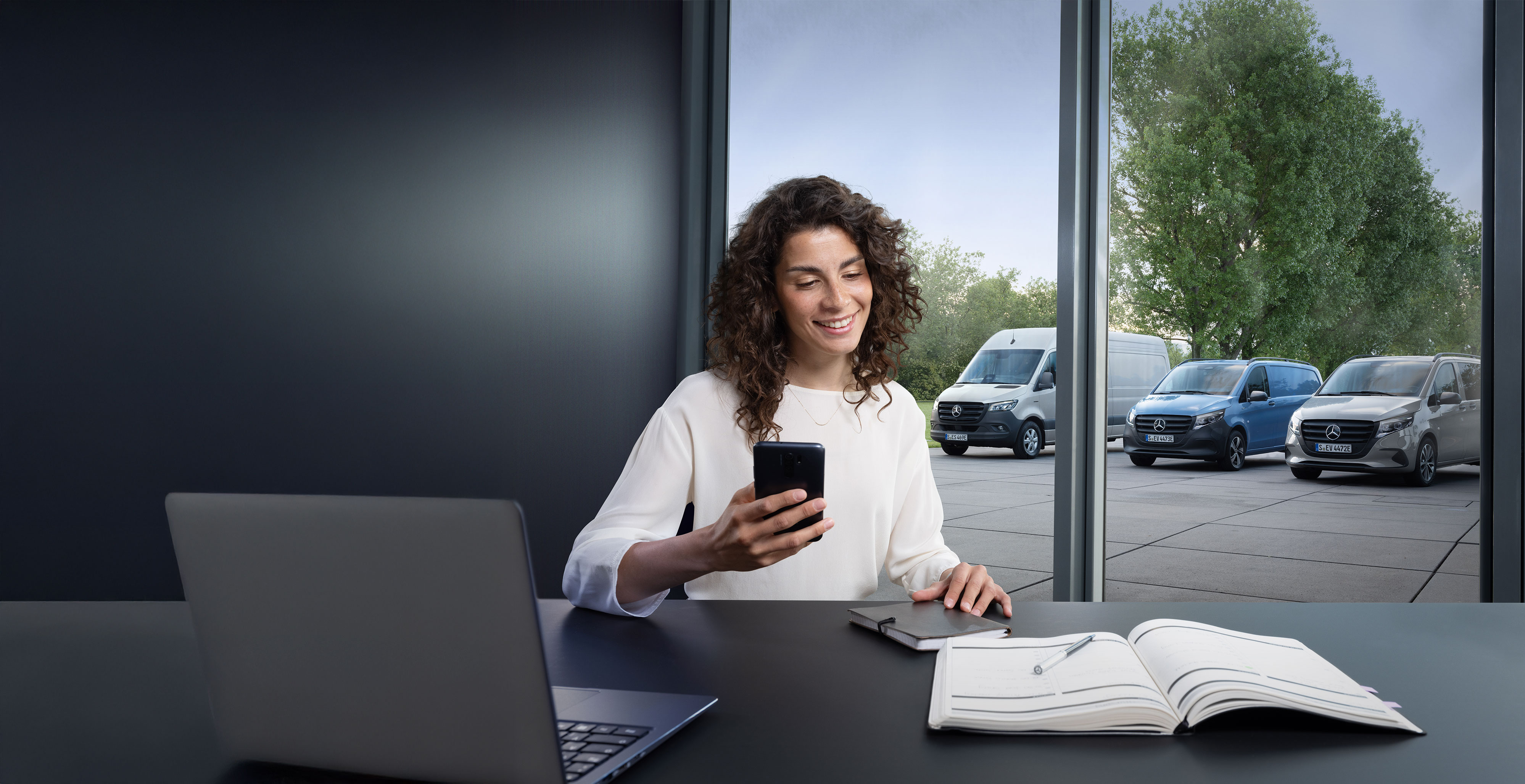 Mercedes-Benz | Hjælp til varebiler Kvinde med smartphone ved et skrivebord, flere Mercedes-Benz varebiler i baggrunden.
