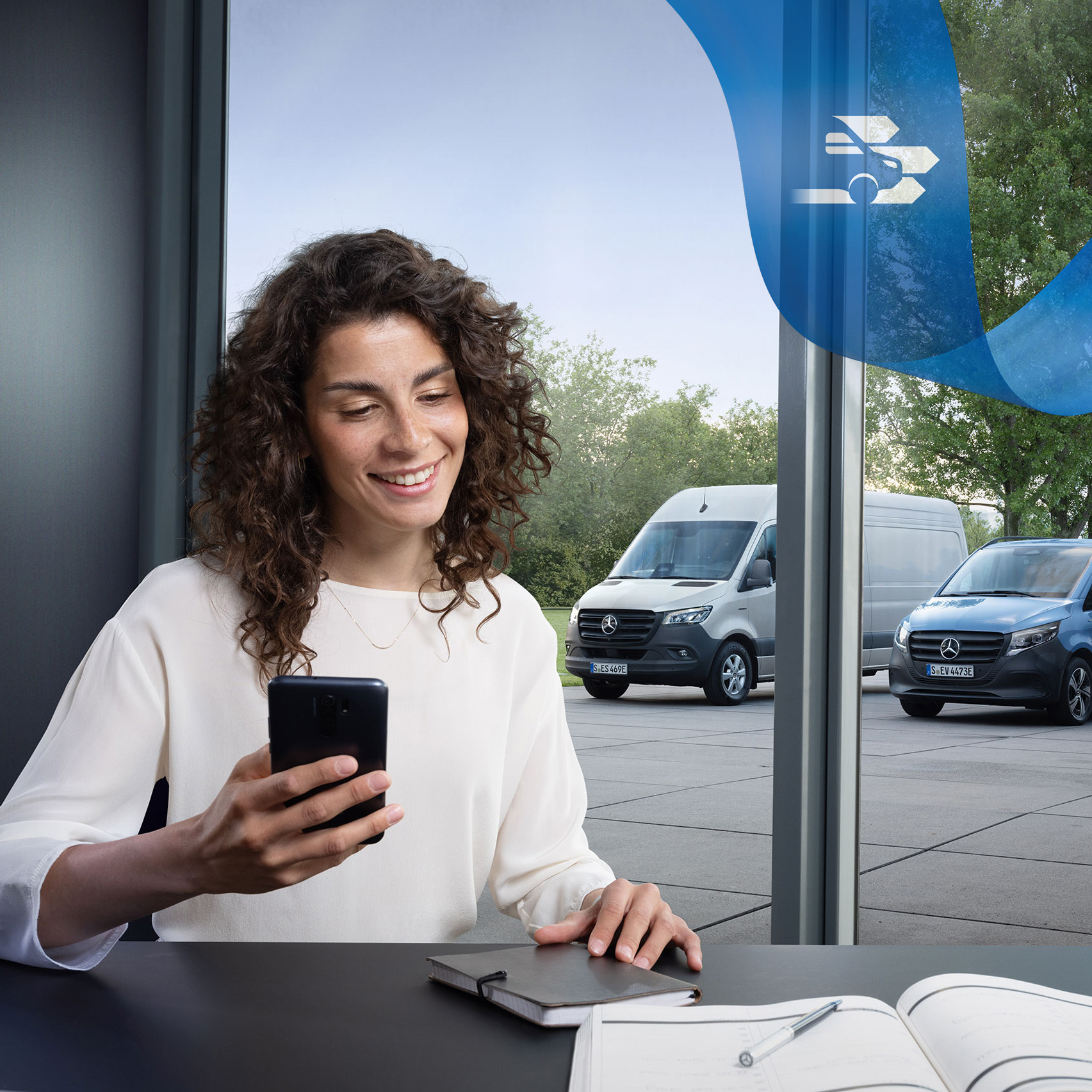 Mercedes-Benz | Varebilsservice | Customer Assistance Center En kvinde sidder ved et skrivebord med en smartphone og et udvalg af Mercedes-Benz varebiler i baggrunden.