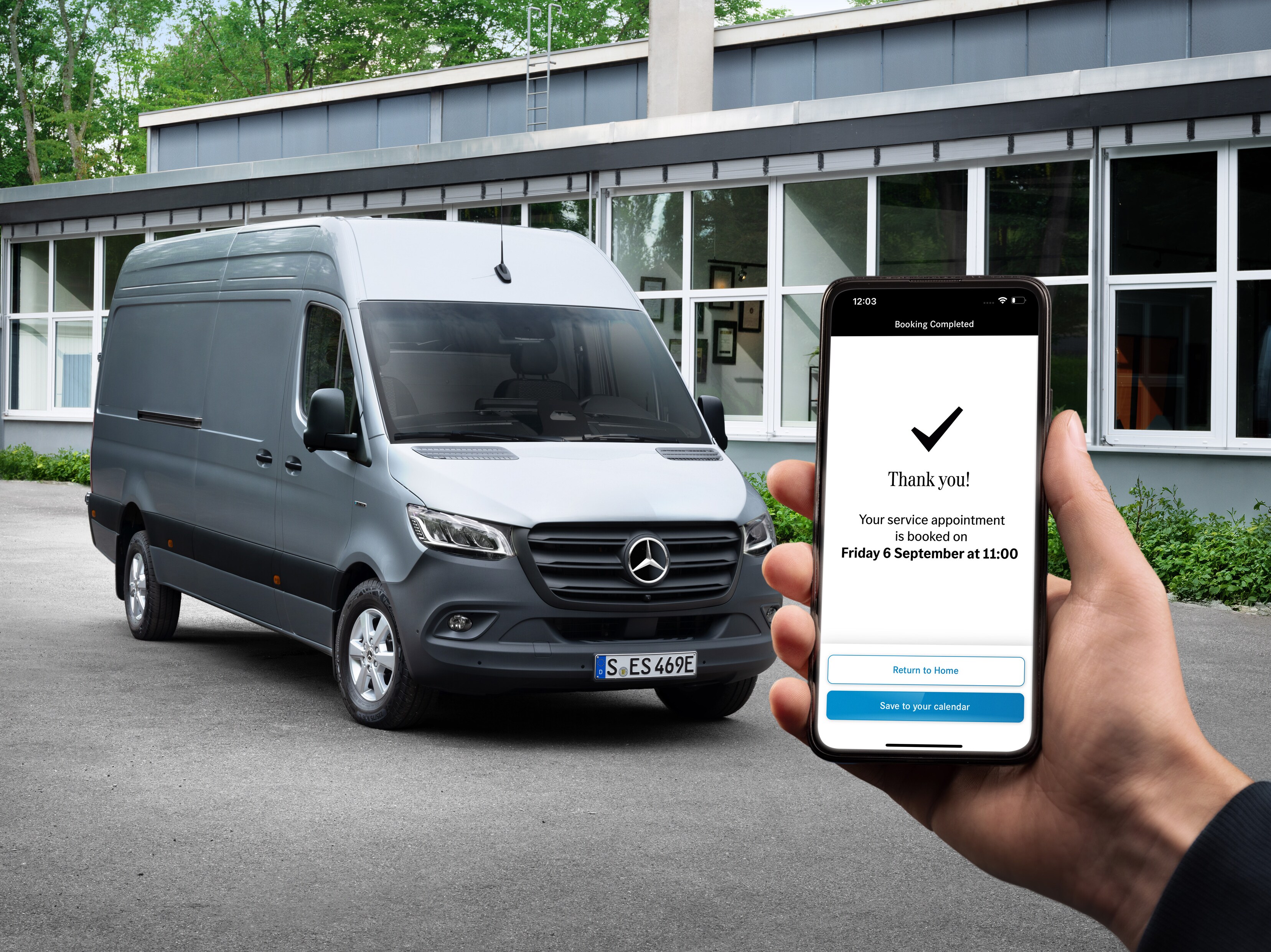 En grå Mercedes-Benz Sprinter varebil foran et kontor med en person, der holder en smartphone i forgrunden.