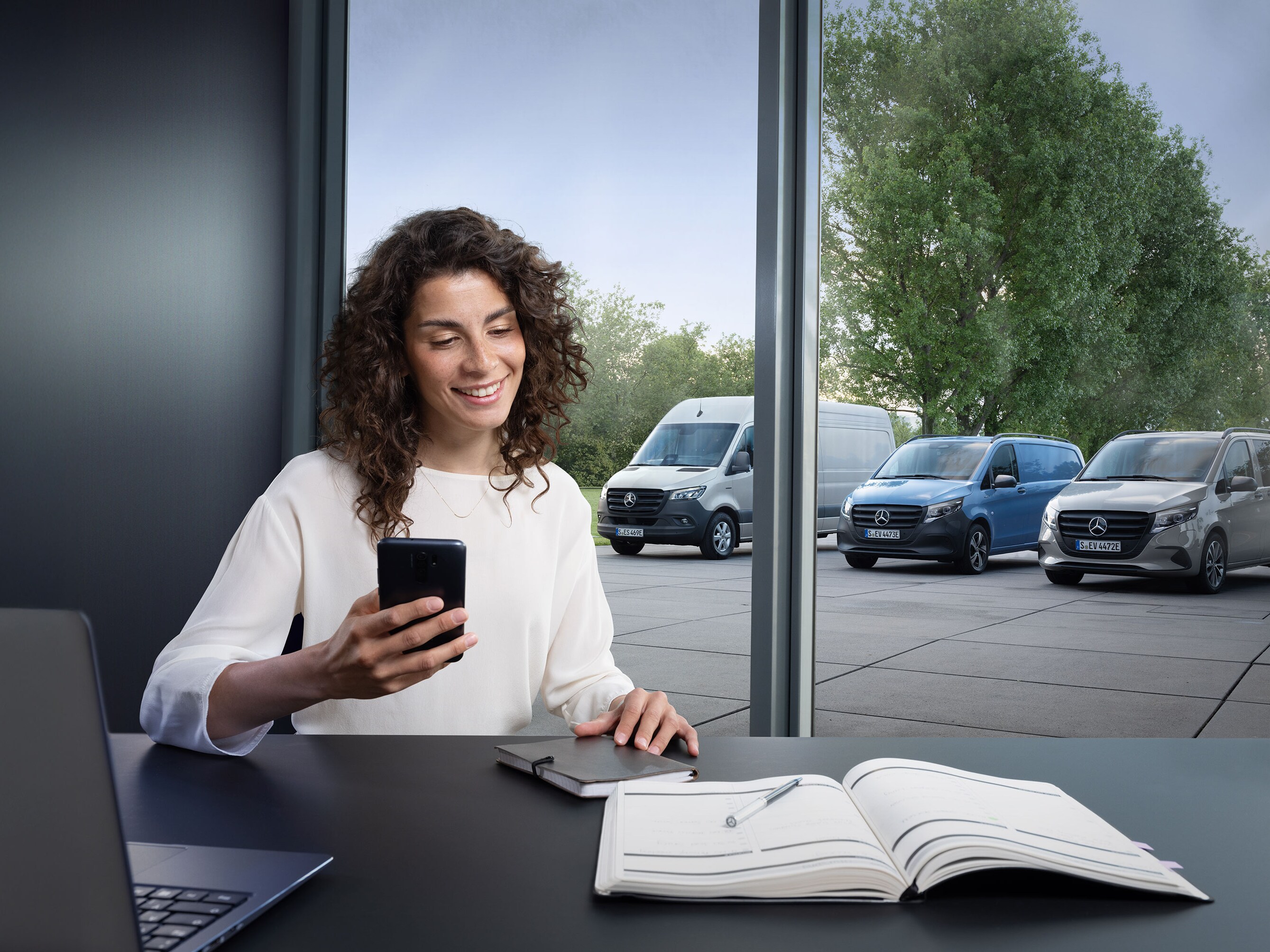 Kvinde booker service via smartphone med en række Mercedes-Benz varebiler i baggrunden.