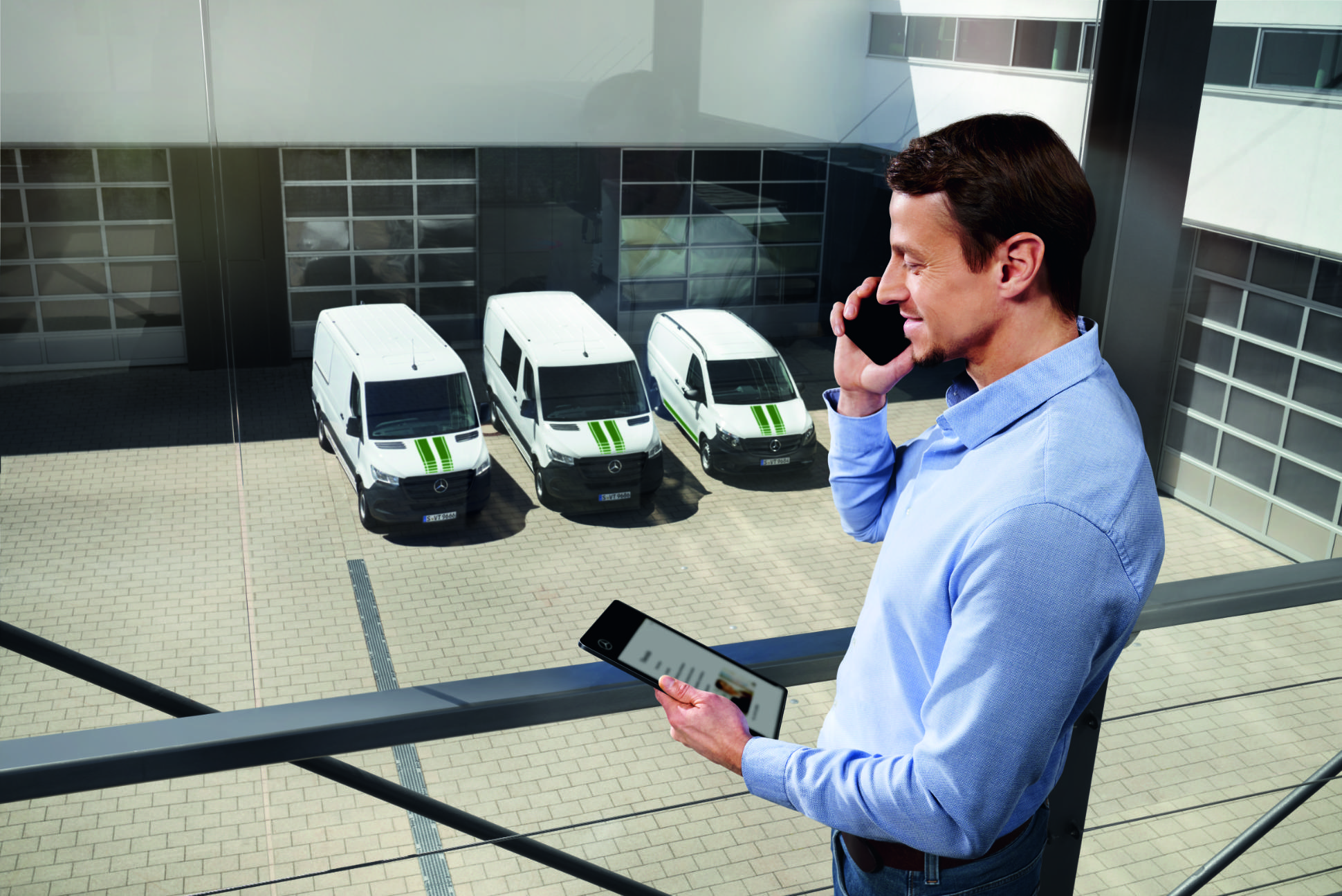 Van Uptime Monitor | Digitale tjenester | Mercedes-Benz Konsulent med en telefon og en tablet i hånden og Mercedes-Benz varebiler i baggrunden.