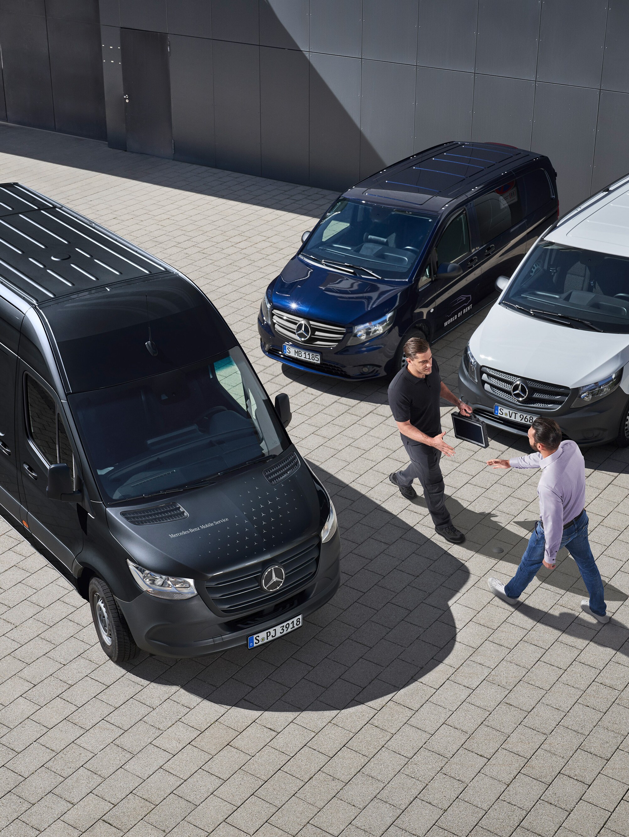 To mænd hilser på hinanden blandt Mercedes-Benz varebiler på en parkeringsplads.