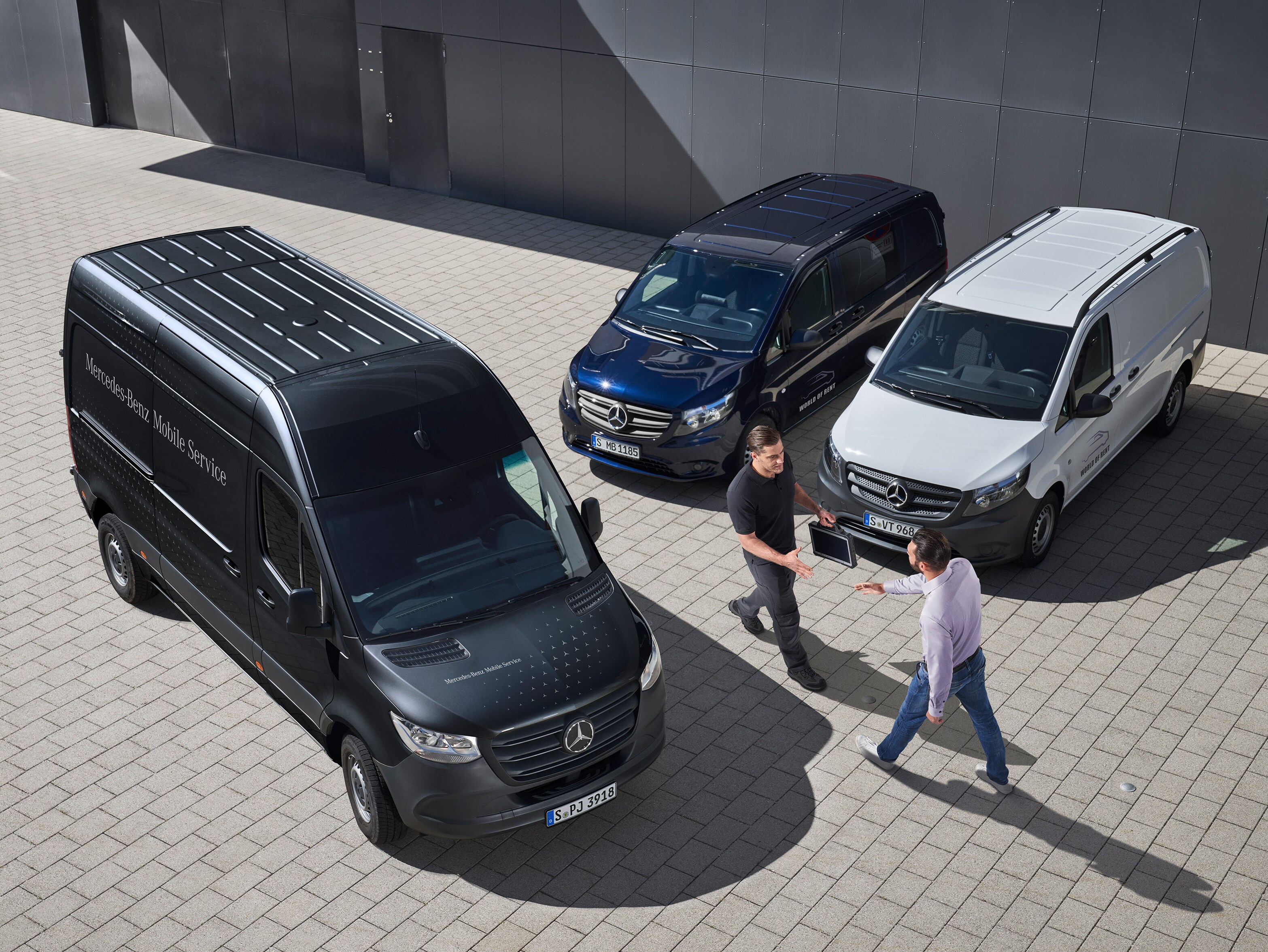 To mænd hilser på hinanden blandt Mercedes-Benz varebiler på en parkeringsplads.