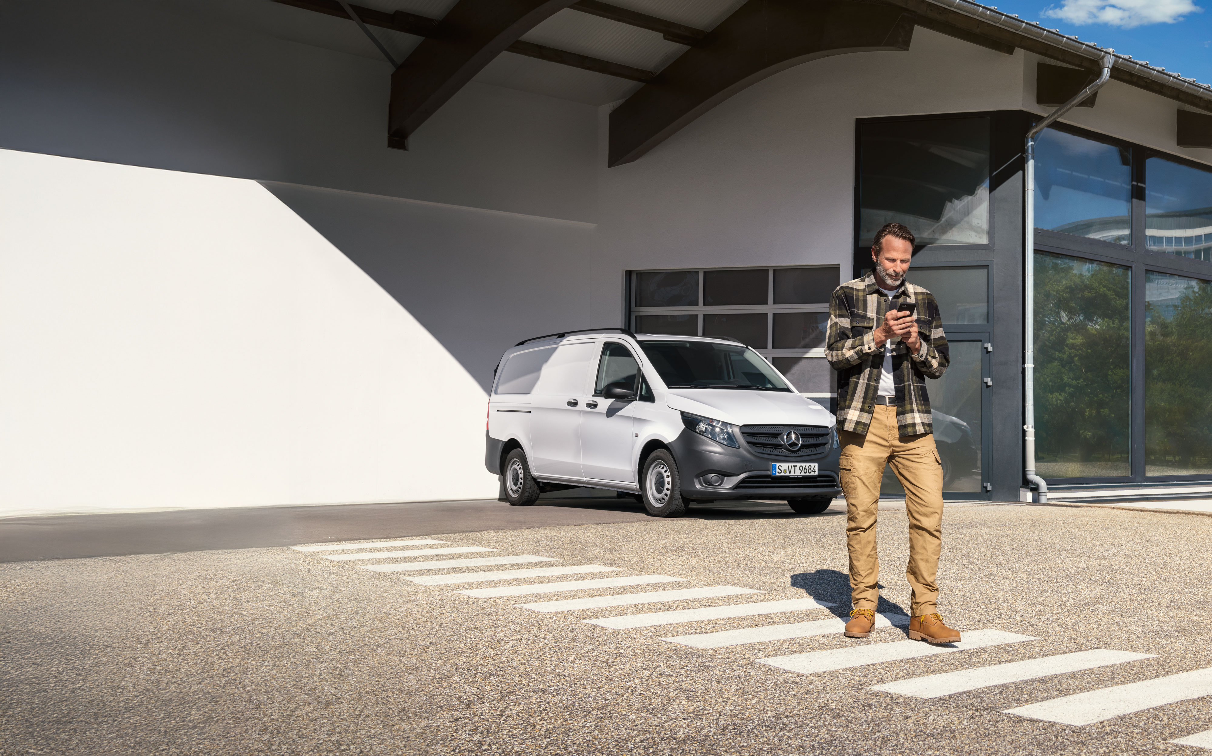 Mercedes-Benz | Bilservice og eftersyn | Servicebooking Kunden booker en aftale på sin telefon, mens han står foran en hvid Mercedes-Benz Vito Varevogn.